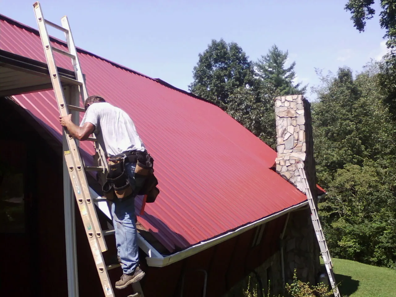 Roofer on a ladder beside a red metal roof and stone chimney — Soffit & Fascia Repair by Roofing Brothers in Blackwell